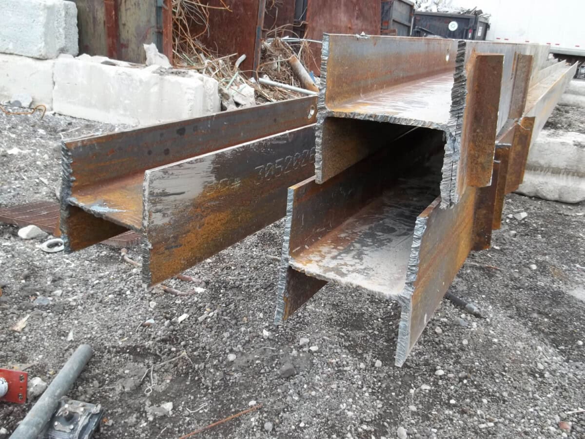 Several large, rusted steel I-beams stacked on gravel, showing rough, freshly cut edges. Concrete debris and metal scraps are scattered in the background.