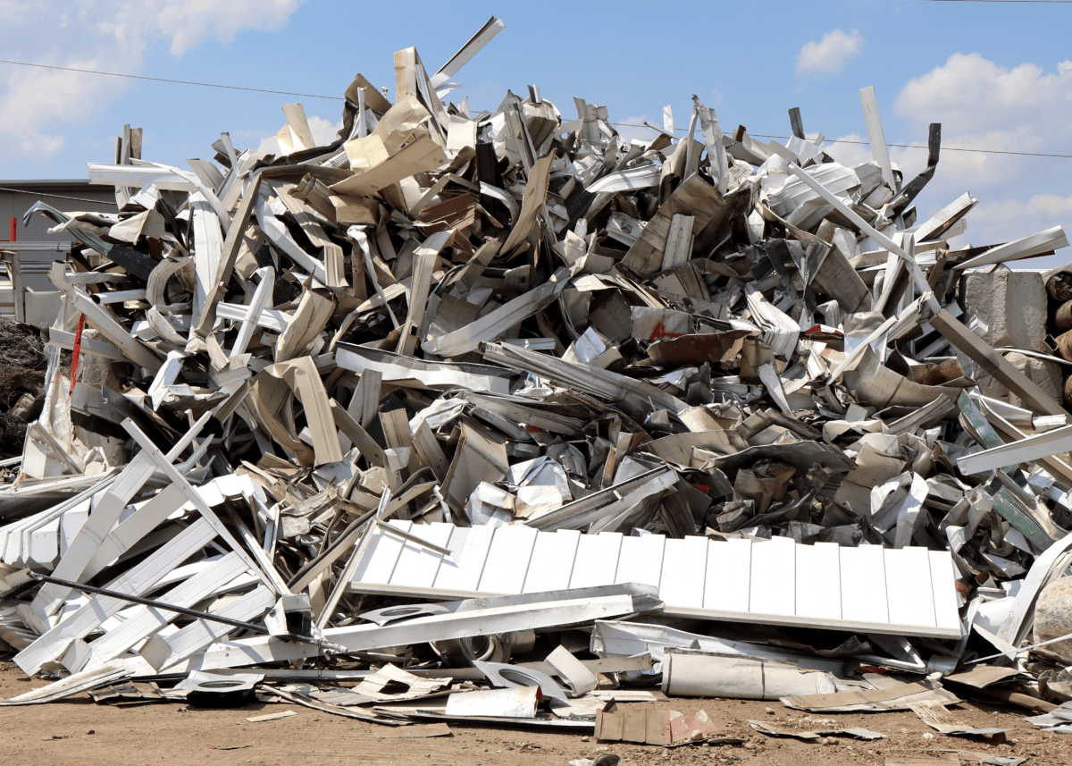 A large pile of assorted metal scraps and debris sits outdoors under a blue sky with clouds, surrounded by dirt and some other materials in the background.