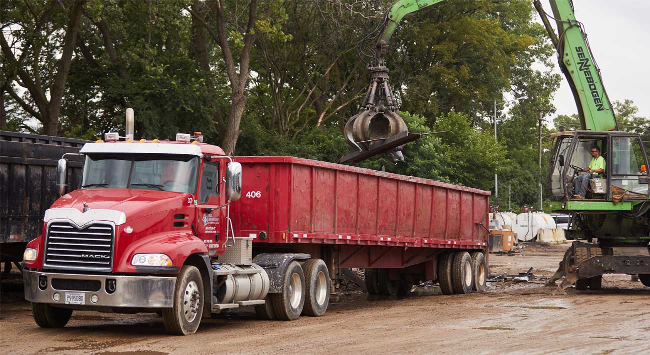 A green excavator loads scrap metal into a large red dump truck at a construction or recycling site with trees in the background.