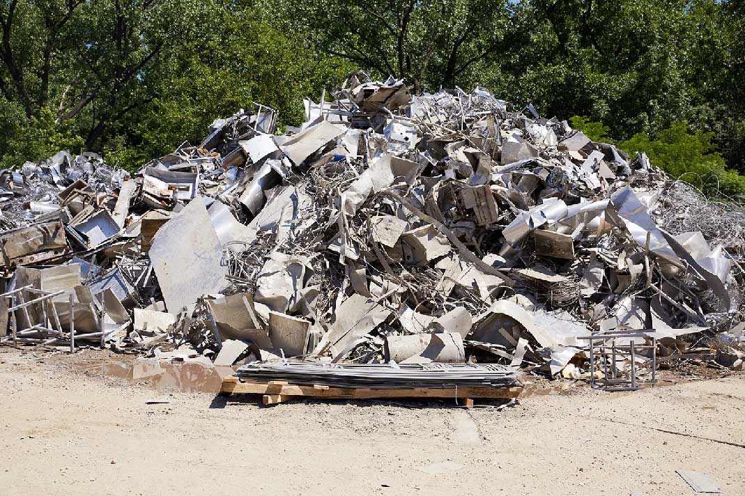 A large pile of scrap metal sits outdoors on bare ground, surrounded by green trees in the background.