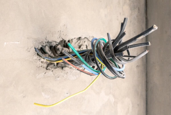 A bundle of exposed electrical wires of various colors protrudes from a hole in an unfinished concrete wall.