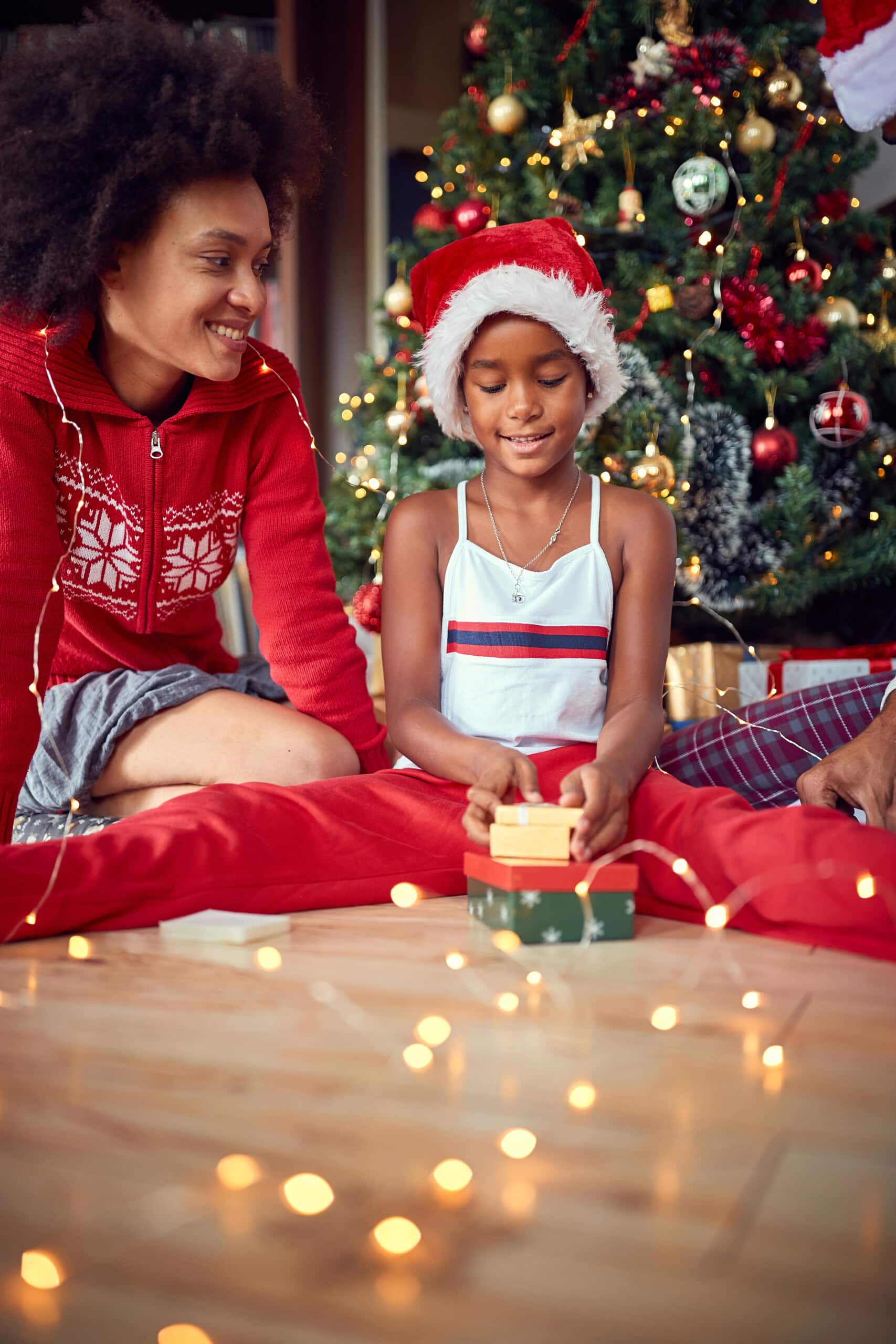 A young child wearing a Santa hat opens a gift box while sitting next to an adult, both smiling, in front of a decorated Christmas tree with festive lights on the floor.