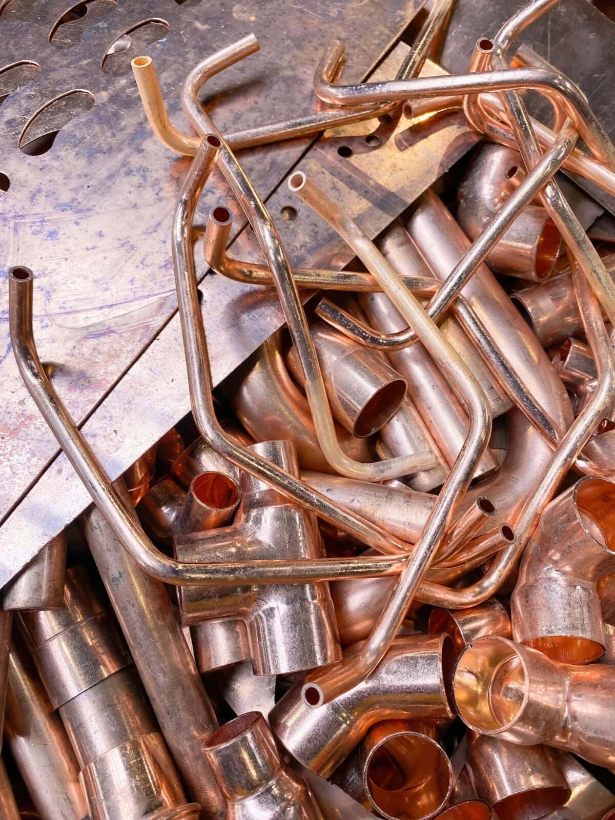 A pile of shiny copper pipes and fittings, some bent and some straight, lying on a metallic surface.