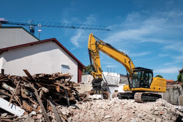 A yellow excavator is demolishing a building, surrounded by piles of rubble and wooden debris, with a blue crane and partially intact structures in the background under a clear sky.