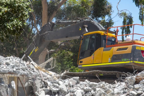 A yellow excavator is clearing rubble and debris at a demolition site, surrounded by trees and blue sky in the background.