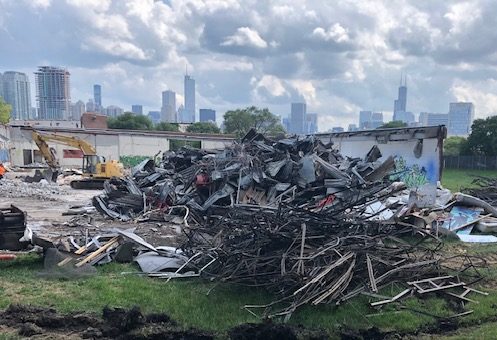 A large pile of debris and twisted metal sits on grass in front of a partially demolished building, with city skyscrapers visible in the background under a cloudy sky.