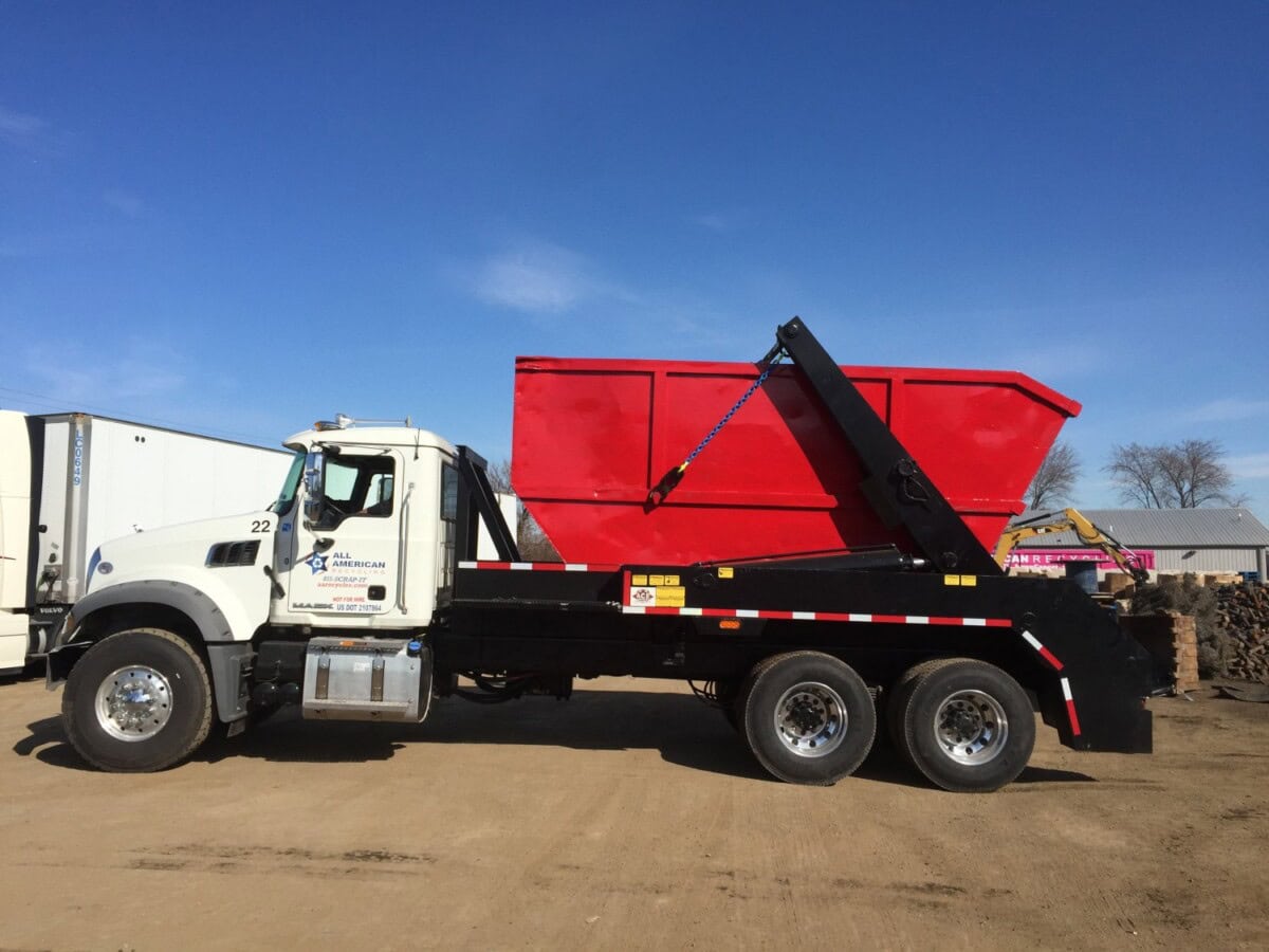 A white truck with black trim is carrying a large, bright red dumpster container on its flatbed. The truck is parked on dirt, with clear blue sky and other vehicles in the background.