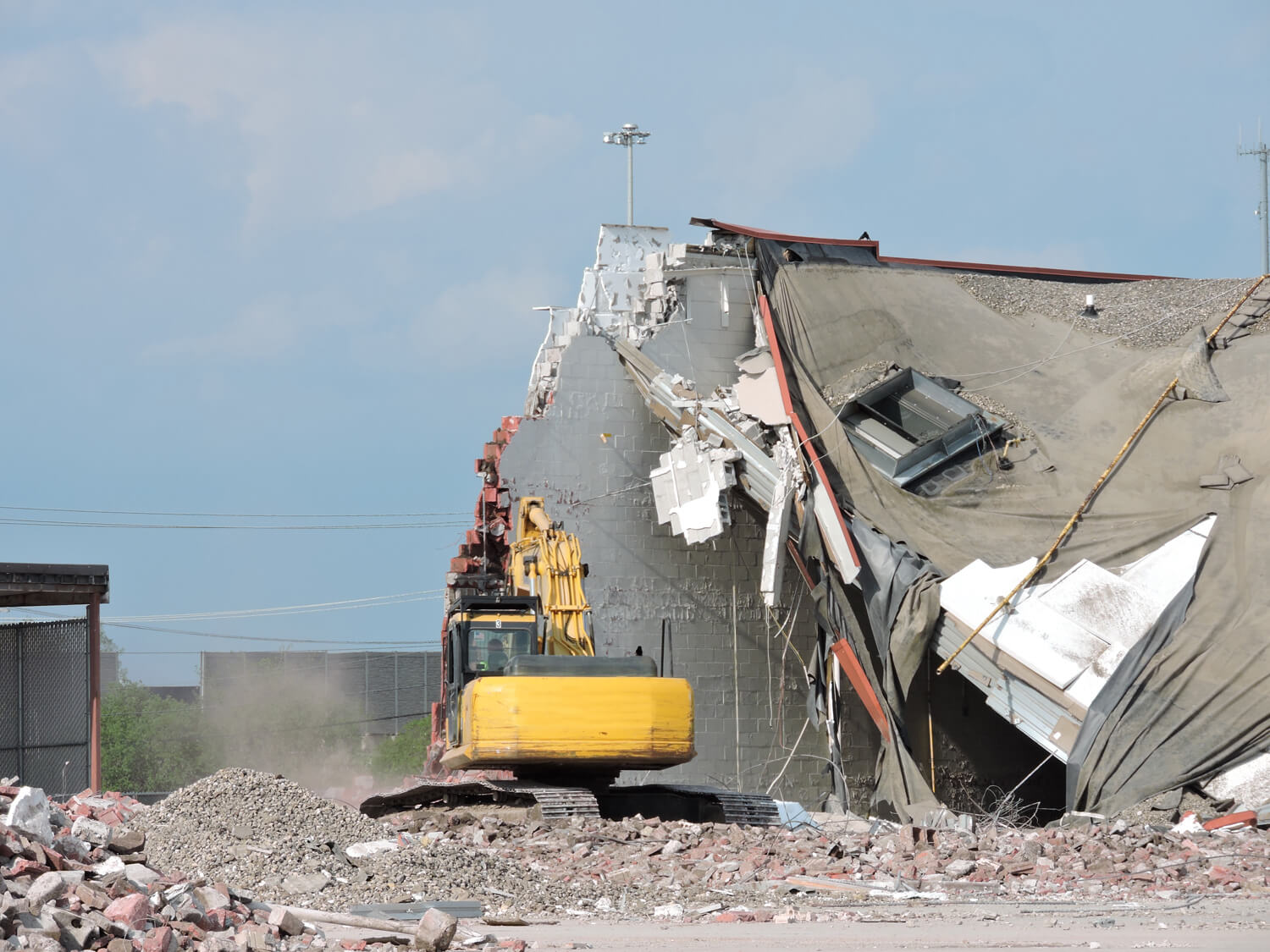 A yellow excavator demolishes a large, partially collapsed building, with rubble and debris scattered around the site under a clear sky.