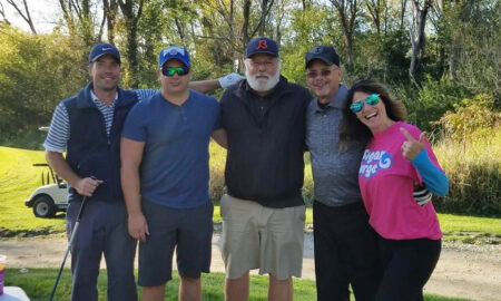 Five people stand together outdoors on a golf course, smiling at the camera. Four men wear casual golf attire; the woman on the right wears a bright pink shirt and sunglasses, giving a thumbs-up gesture. Trees and golf cart are in background.