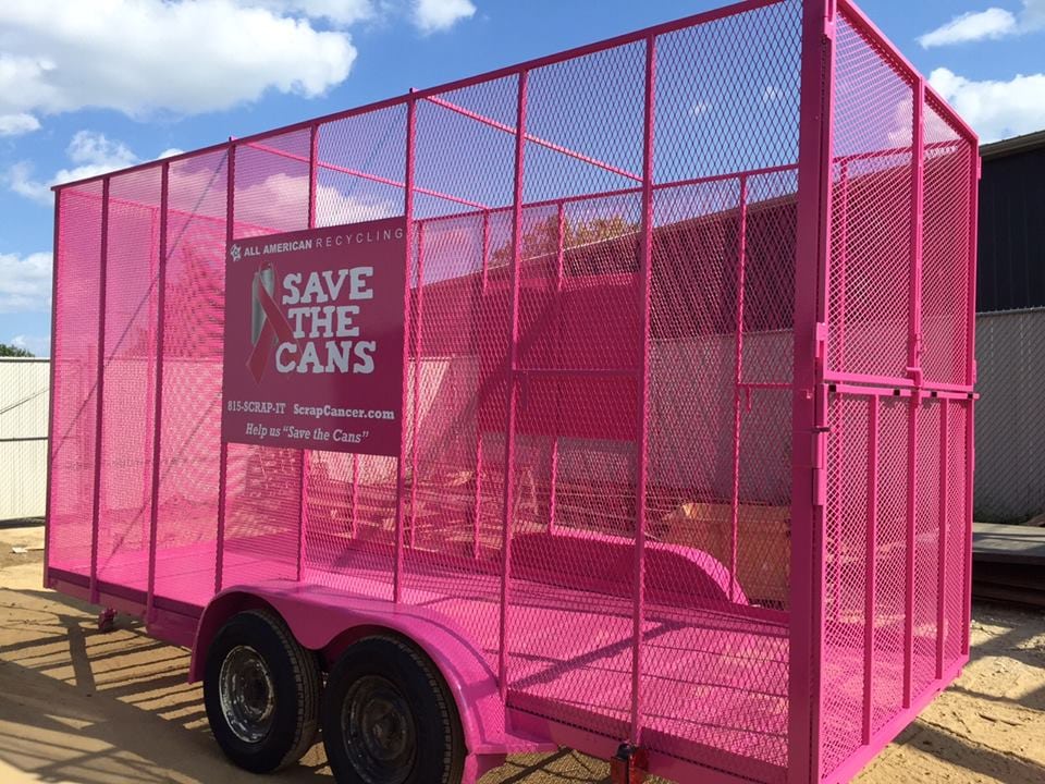 Bright pink metal trailer with mesh sides and a sign reading Save The Cans from All American Recycling. The trailer is parked on dirt near a fence and building, under a blue sky with scattered clouds.