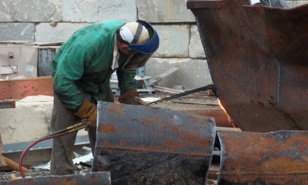 A worker in protective gear uses a torch to cut through large pieces of rusted metal in an outdoor industrial setting with stone walls in the background.