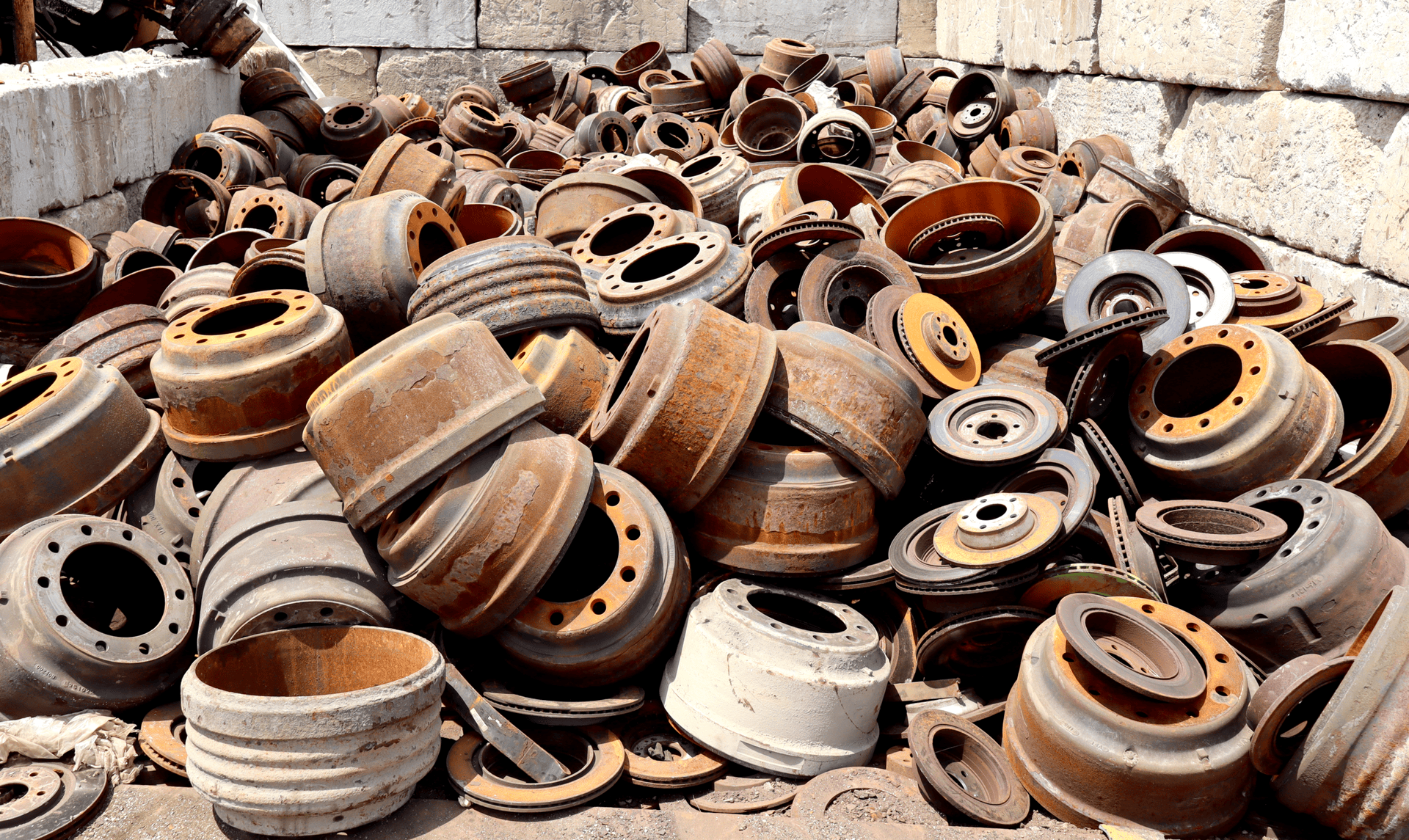 A large pile of rusty, discarded brake drums and car parts stacked against a wall in an open area, suggesting an automotive junkyard or scrap metal collection site.