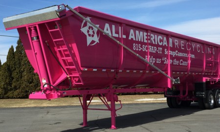 Bright pink recycling trailer with white text, parked on pavement near trees. The side displays a message about helping a cancer cause and features a pink ribbon symbol.