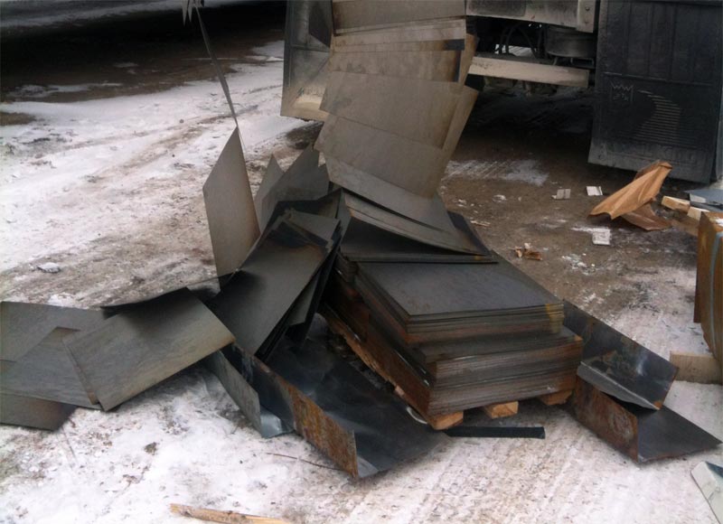 Metal sheets are scattered and stacked unevenly on a snowy, dirty ground near a truck. Some sheets are partially upright while others lay flat, with wooden planks and debris nearby.