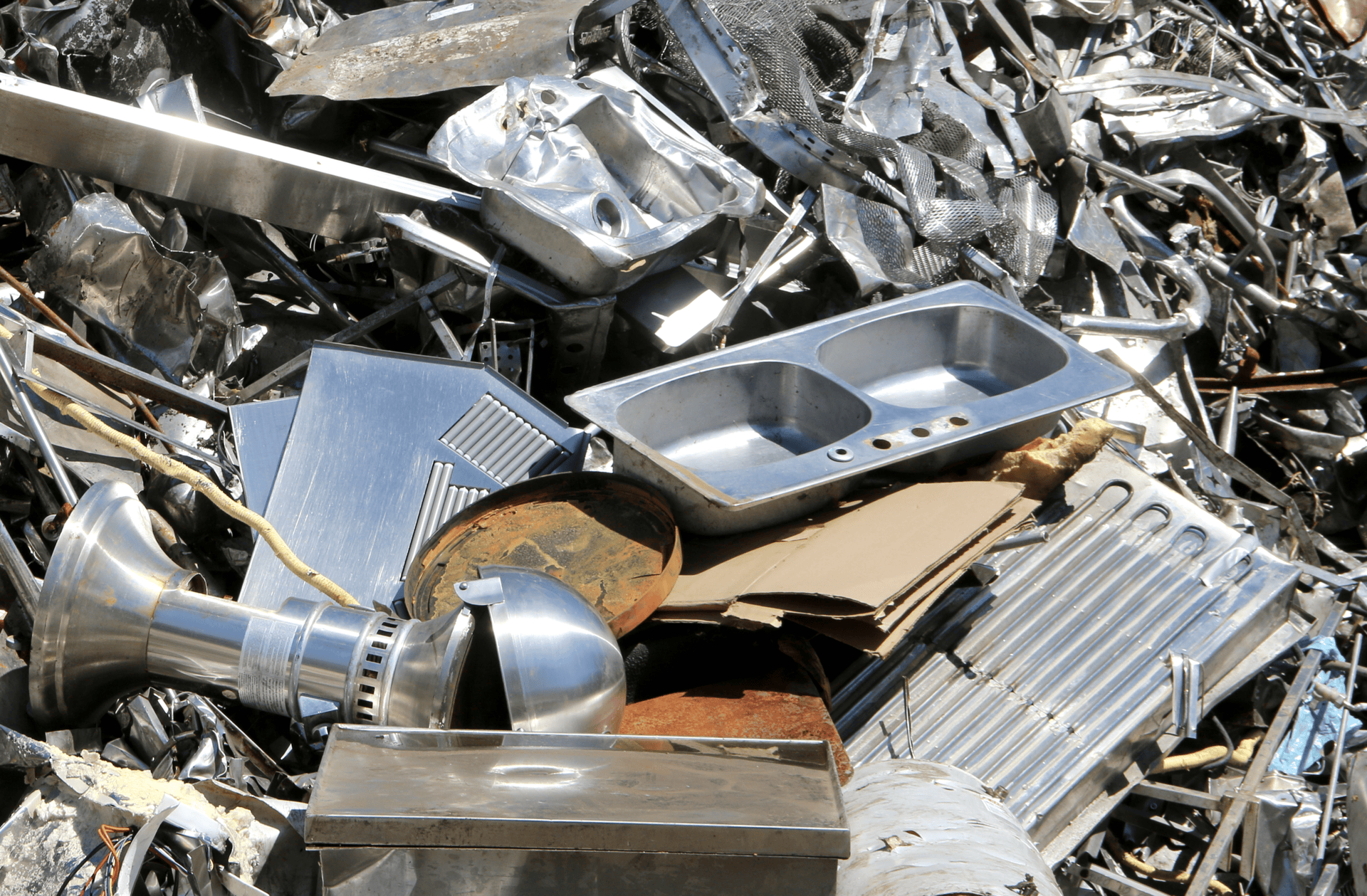 A pile of stainless steel scrap metal, including sinks, industrial equipment, and various metal parts, stacked together in a scrapyard.