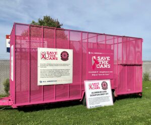A large pink metal donation bin labeled Save the Cans sits on grass, with signs encouraging aluminum can donations for breast cancer awareness.