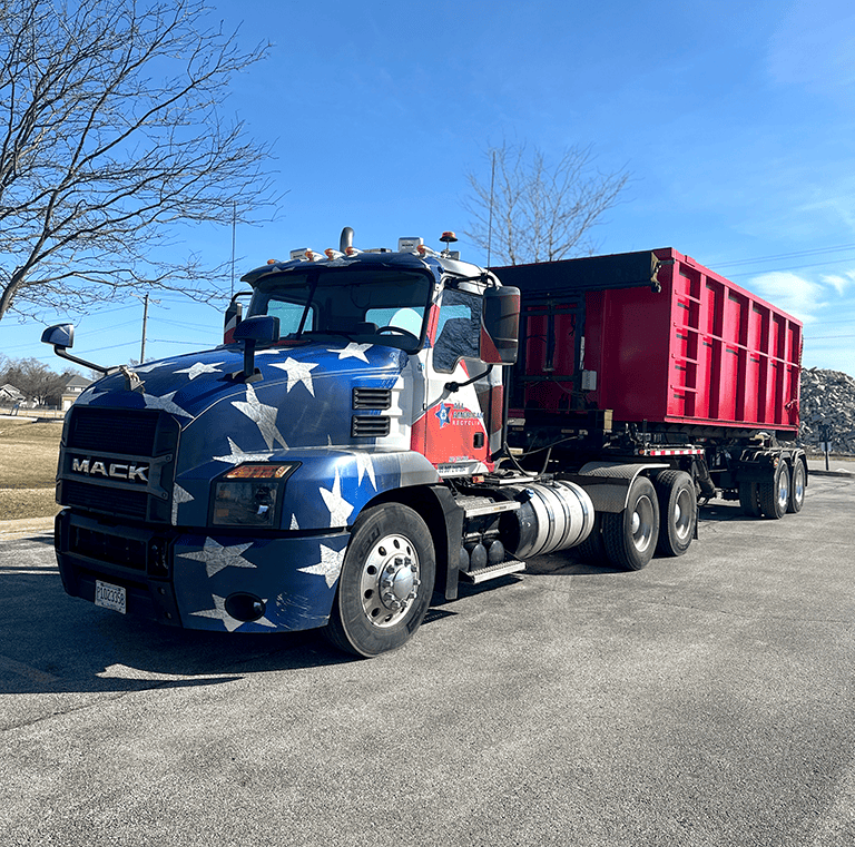 A large Mack truck with an American flag design on the cab and a red dump trailer is parked on a paved lot under a clear blue sky, with bare trees and a rocky area in the background.